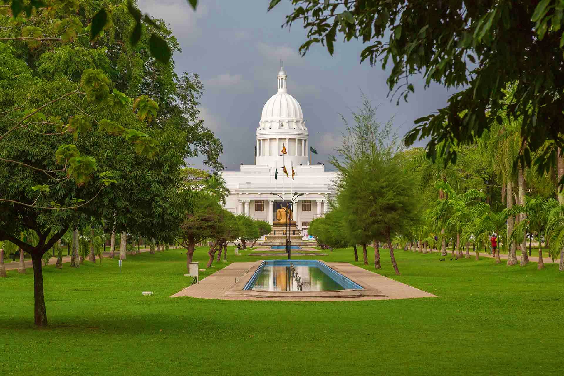 White domed government building viewed through tree-lined park with reflecting pool in foreground, Sri Lanka